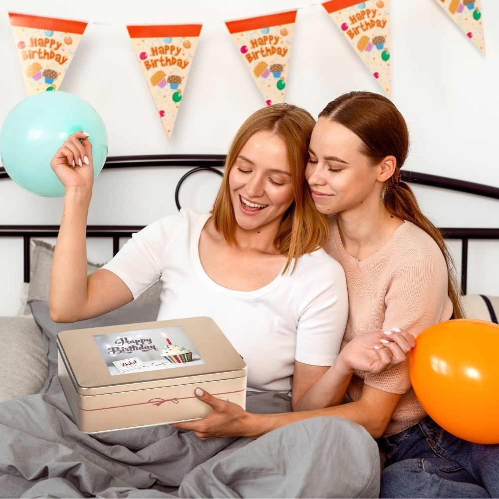 Two smiling women celebrating a birthday with a Dulcet gold gift tin featuring a Happy Birthday cupcake design, colorful balloons, and festive birthday decorations.