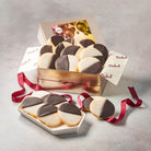 Dulcet gold celebration gift tin filled with chocolate and vanilla iced black and white cookies, featuring a colorful balloon-themed lid, branded tissue paper, and burgundy ribbon.
