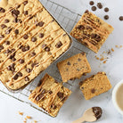 Fresh-baked chocolate chip dessert bars topped with chopped nuts, chocolate chips and a sweet drizzle, with sliced pieces on a cooling rack.