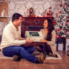 Smiling couple exchanging a Dulcet Happy Holidays gold gift tin beside a decorated Christmas tree and festive fireplace.