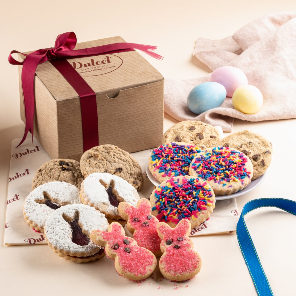 Dulcet Easter bakery gift box tied with a burgundy ribbon, surrounded by pink bunny sugar cookies, bunny Linzer cookies, rainbow sprinkle cookies, chocolate chip cookies, oatmeal raisin cookies and pastel Easter eggs.