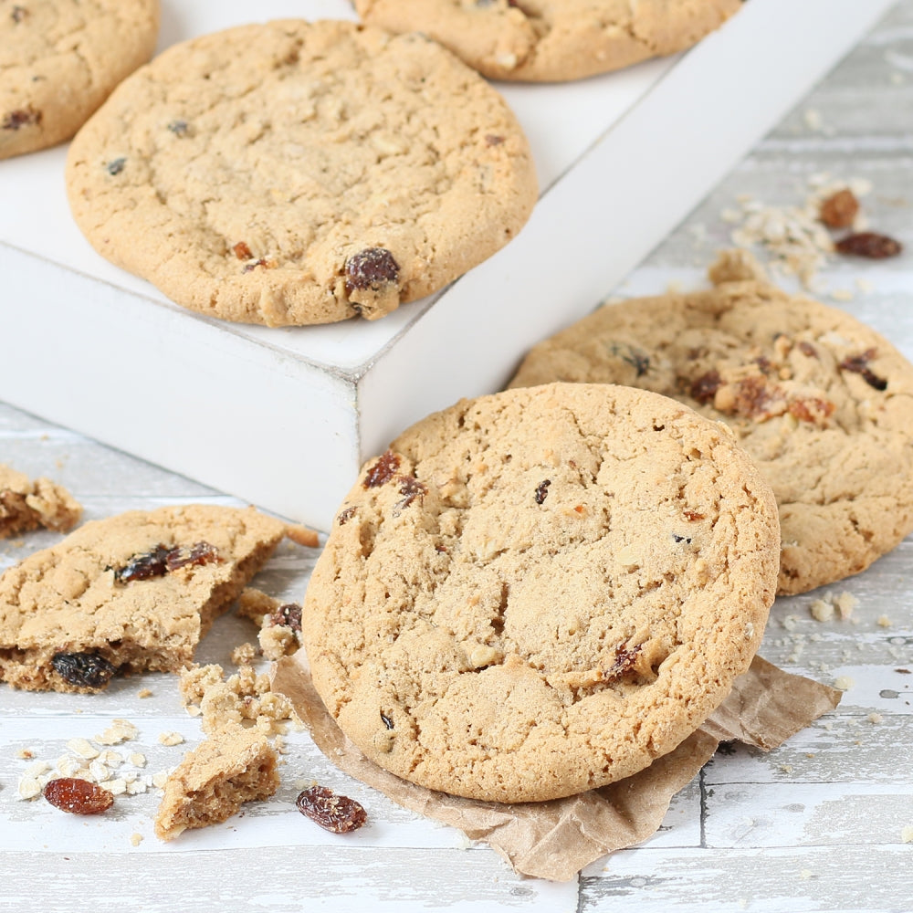 Close-up of Dulcet golden-baked oatmeal raisin cookies with visible raisins and oats, displayed on a rustic white wooden surface with broken cookie pieces.