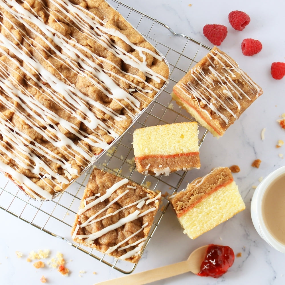 Dulcet raspberry crumb cake with golden streusel topping, white icing drizzle and raspberry filling, shown sliced on a cooling rack with fresh raspberries.