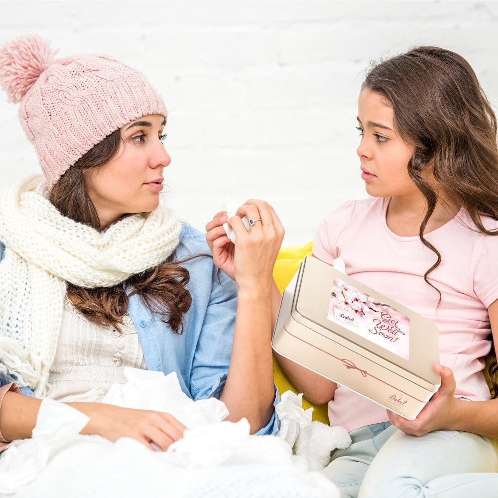 Young girl giving a Dulcet Get Well Soon gourmet gift tin to a woman resting under a blanket, showing a thoughtful gesture of care and support.