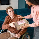 Smiling man in a wheelchair receiving a Dulcet Get Well Soon gold gift tin from a woman, sharing a thoughtful gifting moment at home.