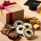 Dulcet graduation gift box with decorated graduation cap cookies, assorted brownies, blondies and rugelach beside a graduation cap.