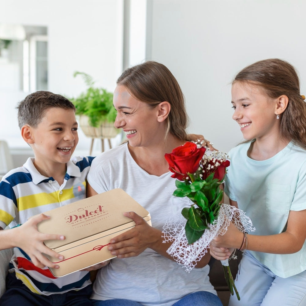 Mother smiling with her son and daughter as they present a Dulcet gourmet gift tin and a bouquet of red roses for a special family celebration.