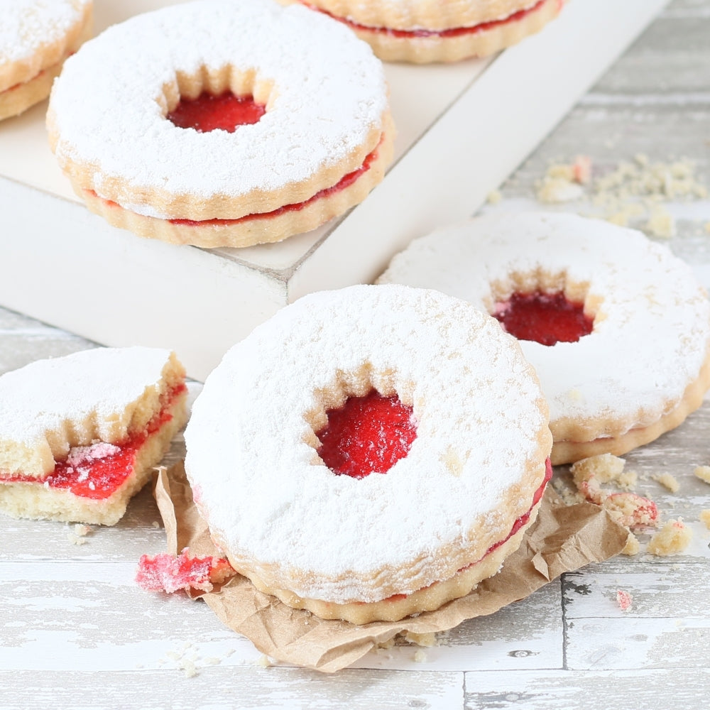 Close-up of raspberry jam-filled Linzer sandwich cookies dusted with powdered sugar, featuring golden buttery cookie layers and red fruit filling.