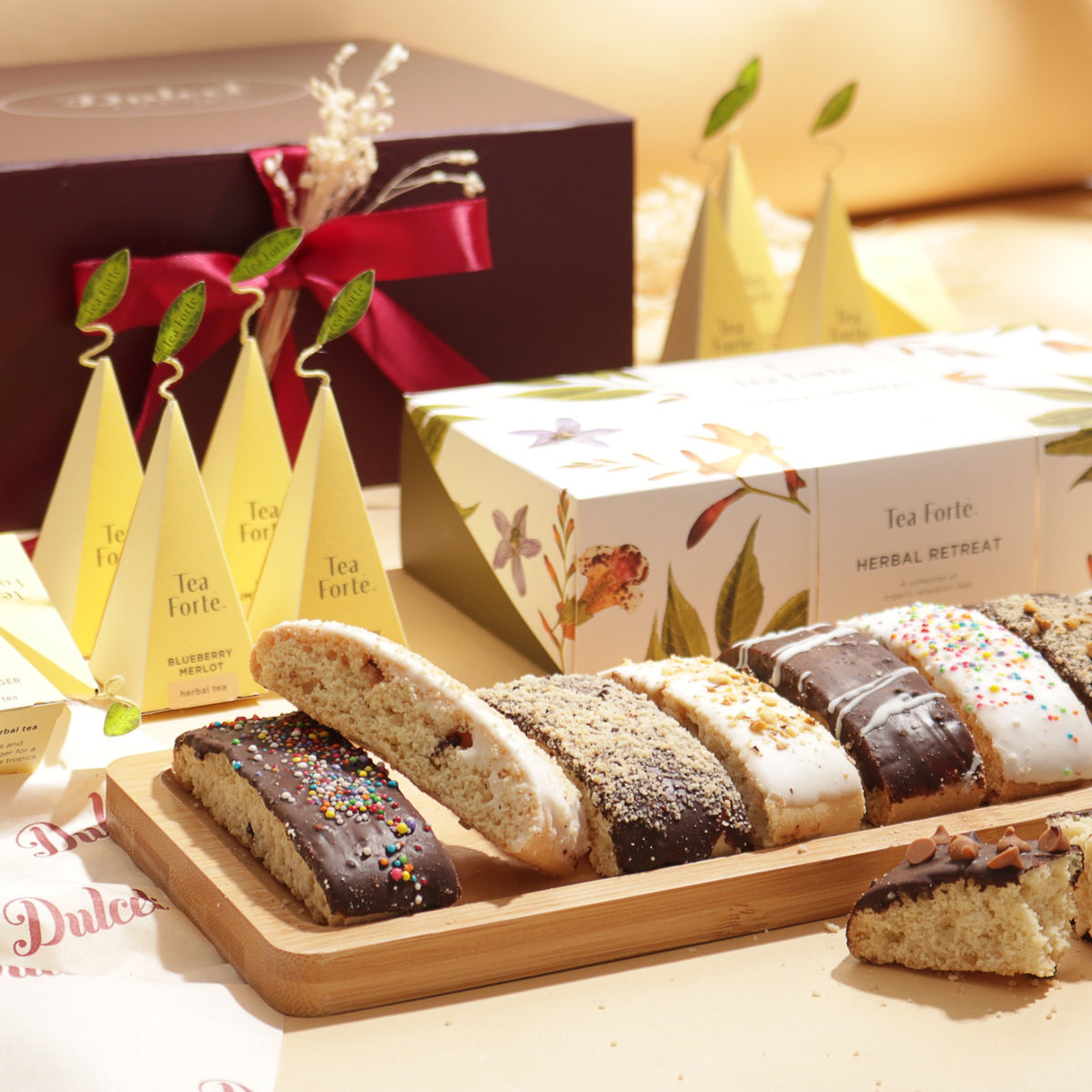 Tea Forté Herbal Retreat tea gift box with pyramid tea infusers, assorted chocolate-dipped biscotti on a wooden tray and a burgundy gift box with red ribbon.