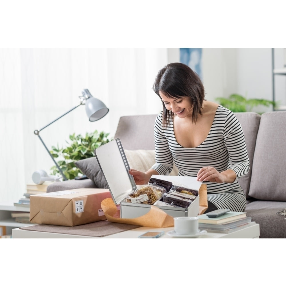 Smiling woman opening a Dulcet gourmet gift tin filled with individually wrapped brownies and bakery treats, with a delivery box beside her at home.