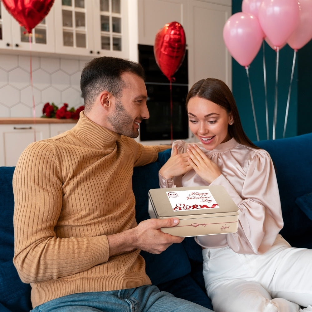Man surprising a smiling woman with a Dulcet Valentine's Day gold gift tin featuring a romantic heart-themed lid, surrounded by pink and red balloons.