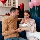 Man surprising a smiling woman with a Dulcet Valentine's Day gold gift tin featuring a romantic heart-themed lid, surrounded by pink and red balloons.