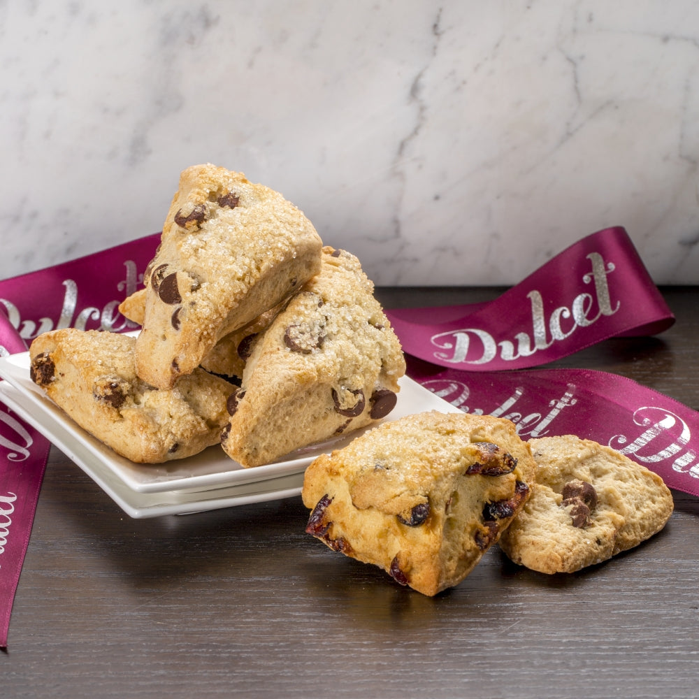 Chocolate chip scones piled on a white plate and dark tabletop with a burgundy Dulcet ribbon.