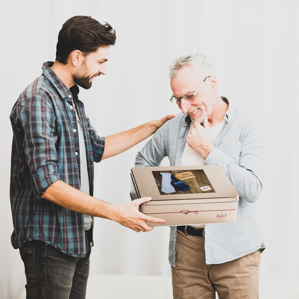 A younger man presents a Dulcet Father’s Day gold gift tin to a smiling older man.