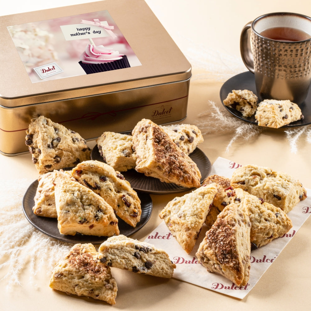 Assorted scones displayed beside a Dulcet gold Mother’s Day gift tin with a pink cupcake design and a mug of tea.