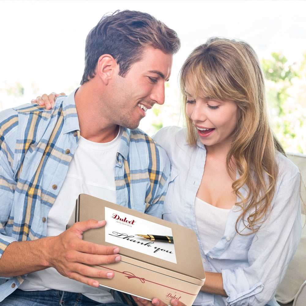 A smiling couple holding a Dulcet thank-you gift tin with a fountain pen design on the lid.