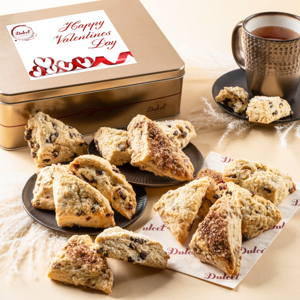 Assorted scones displayed with a Dulcet gold Valentine’s Day gift tin, red ribbon lid design and a mug of tea.