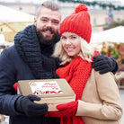 A smiling couple in winter clothing holding a Dulcet gold Valentine’s Day gift tin outdoors.